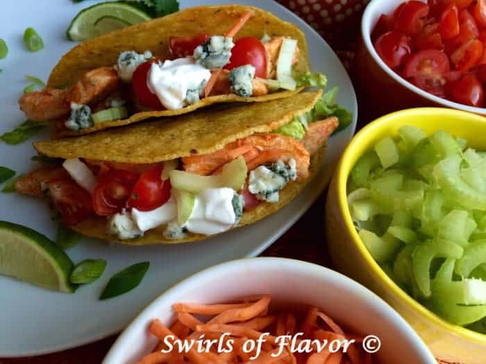Buffalo chicken tacos on a white plate next to bowls containing toppings like tomatoes and celery.