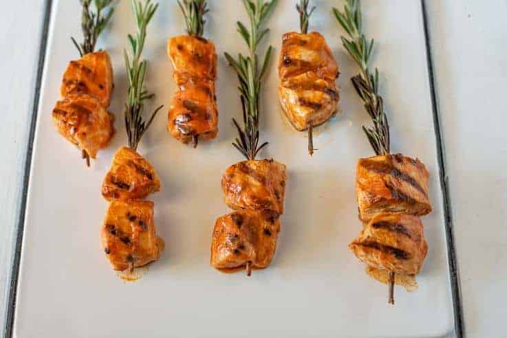 Six pieces of buffalo chicken on rosemary skewers resting on a plate.