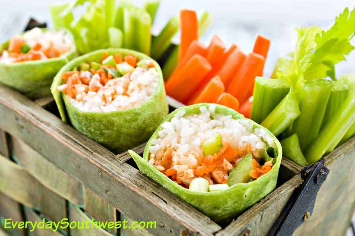 Three buffalo chicken burritos placed upright in a wooden basket next to celery sticks and carrots.