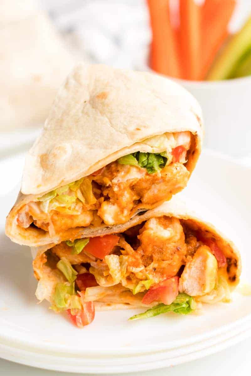 Two buffalo chicken wraps resting on a white plate with carrot and celery sticks in the background.