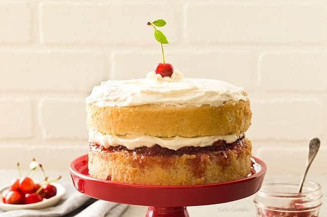 A side view of a cherry vanilla layer cake on a red serving platter with cherries on a dish in the background.