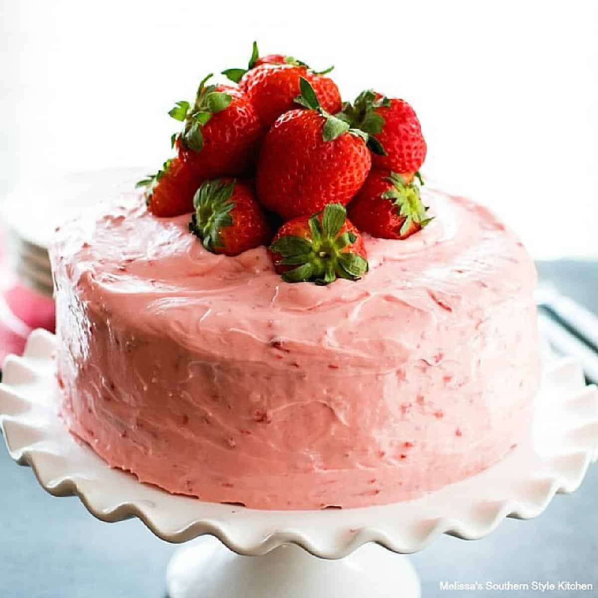 A side view of a strawberry layer cake topped with fresh strawberries resting on a white cake stand.