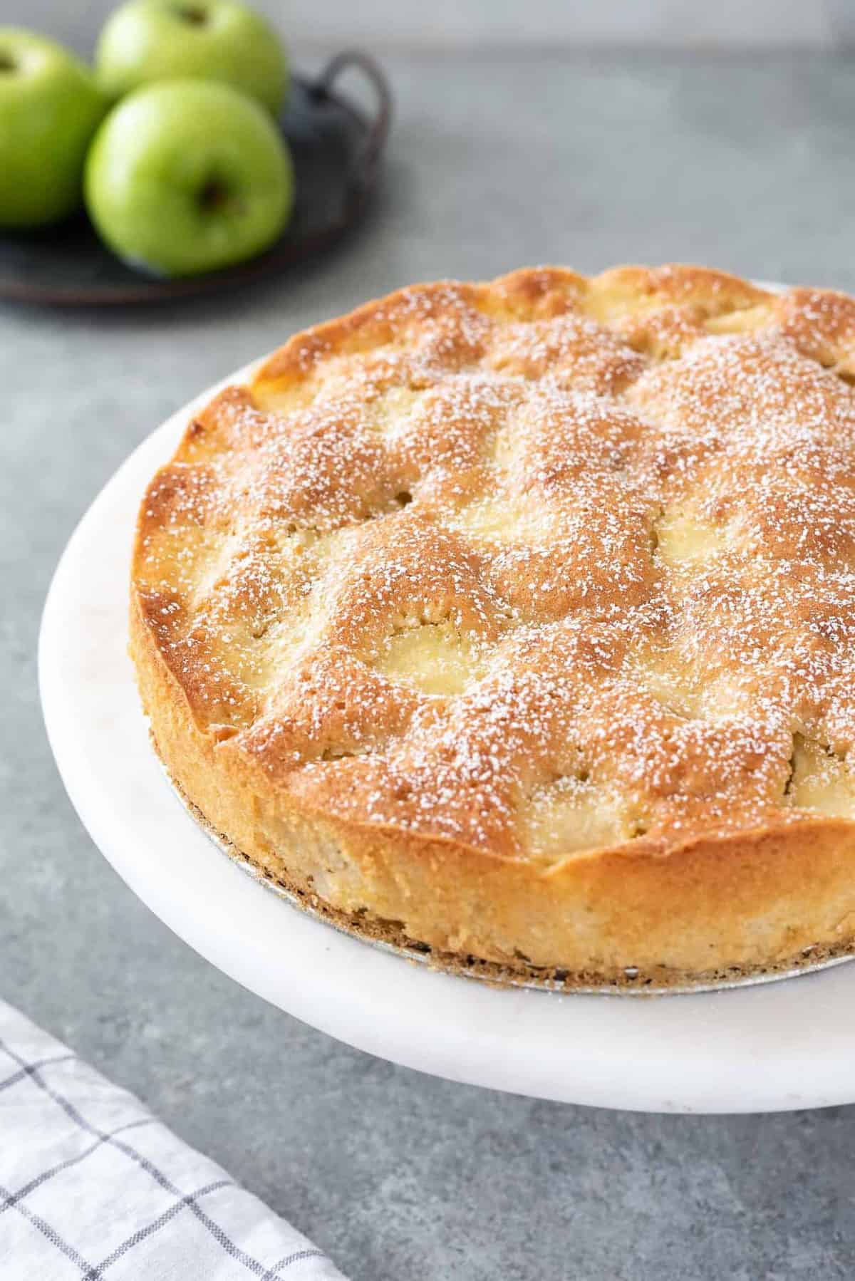 A close up view of French apple cake on a white plate with green apples in the background.