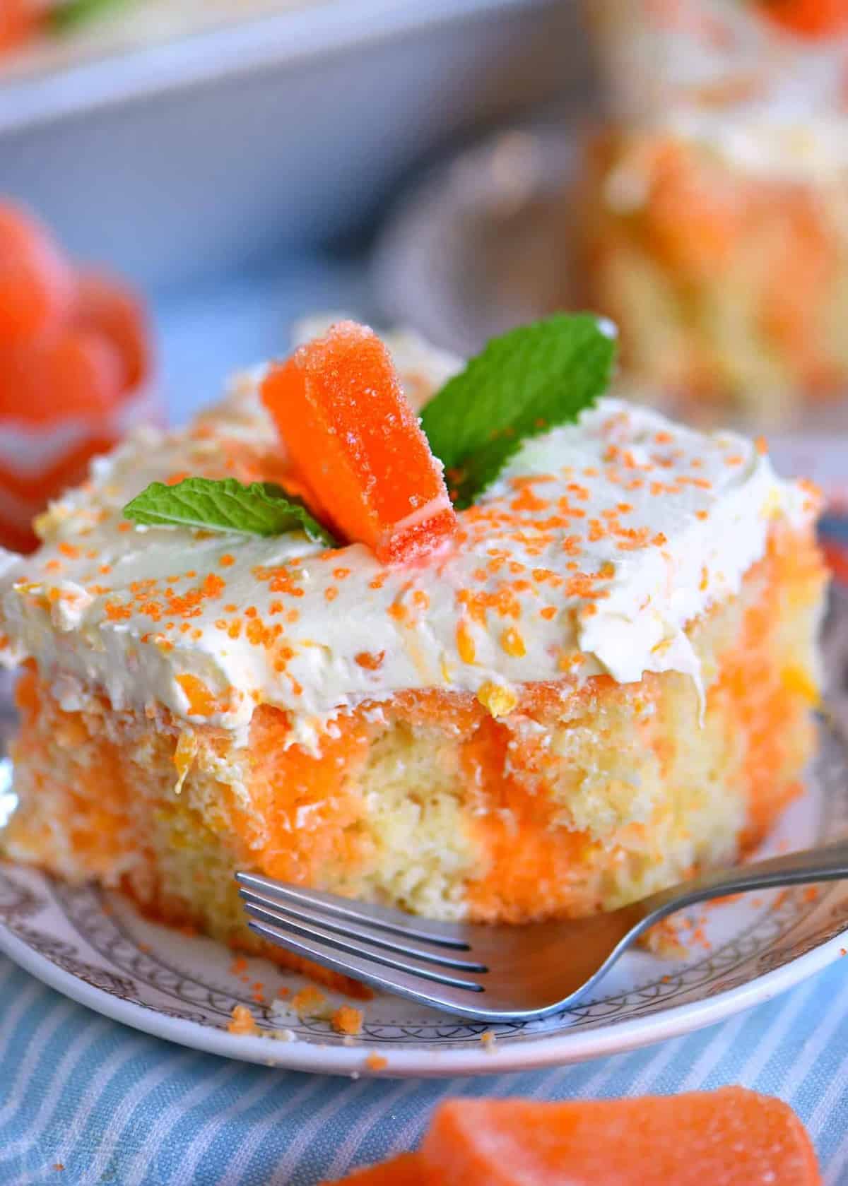 A close up side view of a slice of orange creamsicle cake with mandarin orange frosting on a plate next to a fork.
