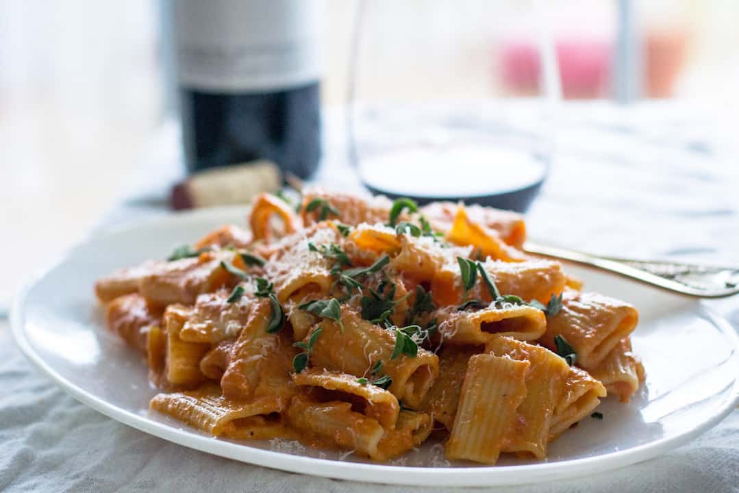 A close up view of penne pasta with a pink vodka sauce on a white plate with a bottle of wine in the background.