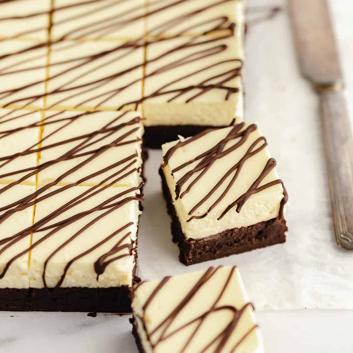 A close up top down view of slices cheesecake brownies on parchment paper next to a butter knife.
