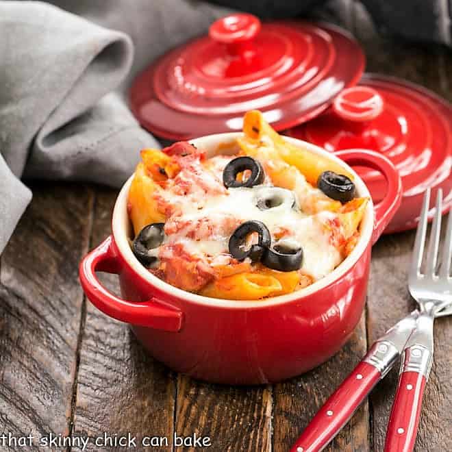 A close up view of cheesy pasta with black olives in a small ceramic bowl on a wooden table.