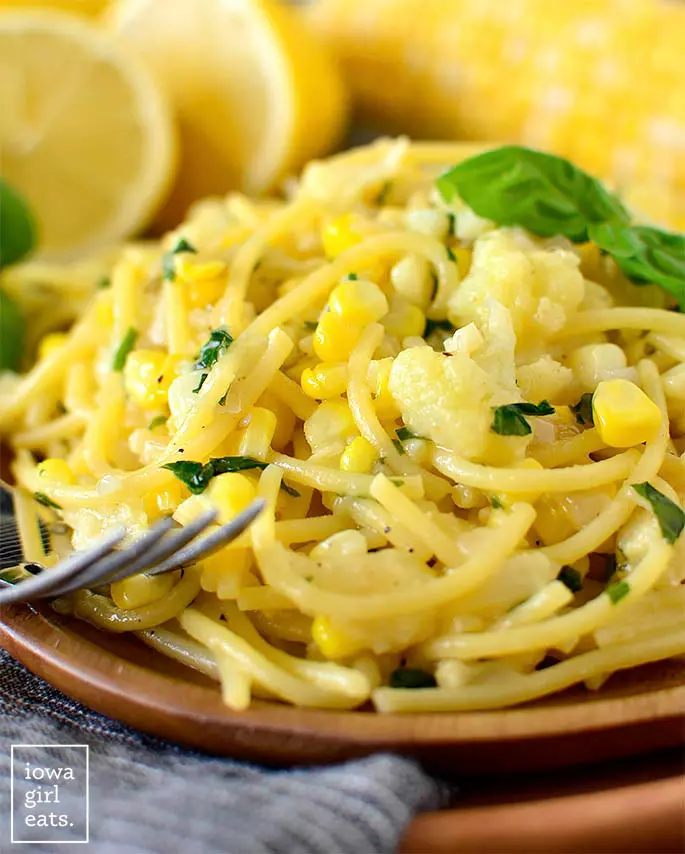 A close up view of creamy sweet corn pasta on a wooden plate with corn and fresh lemon in the background.