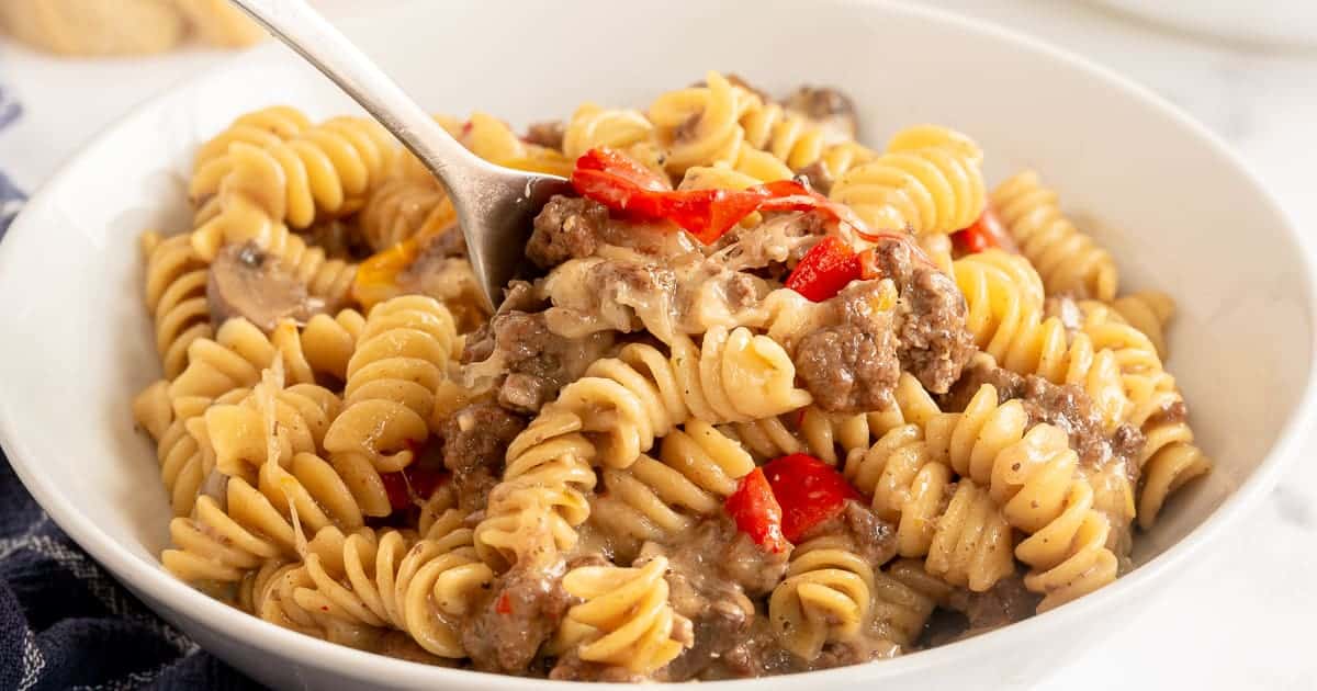 A close up view of Philly cheesesteak pasta in a white bowl with a fork.