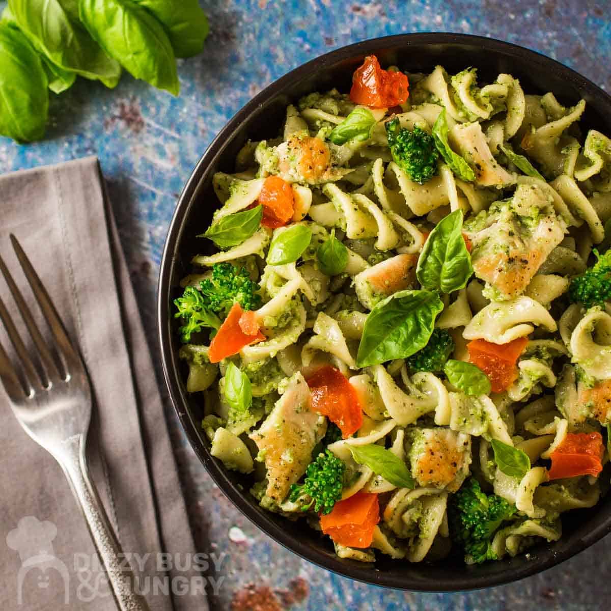 A top down view of chicken pesto pasta on a black plate next to a table setting.