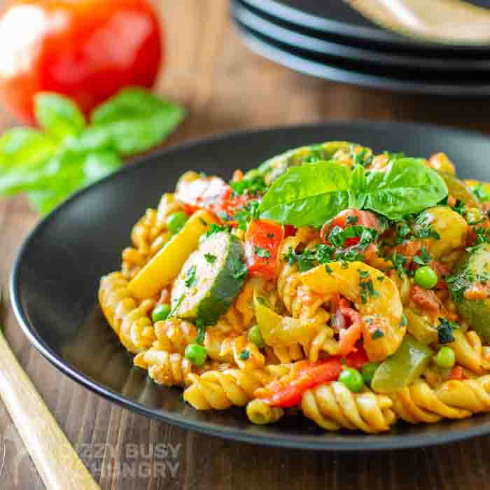 A close up view of curry pasta and vegetables on a black plate resting on a wooden table.