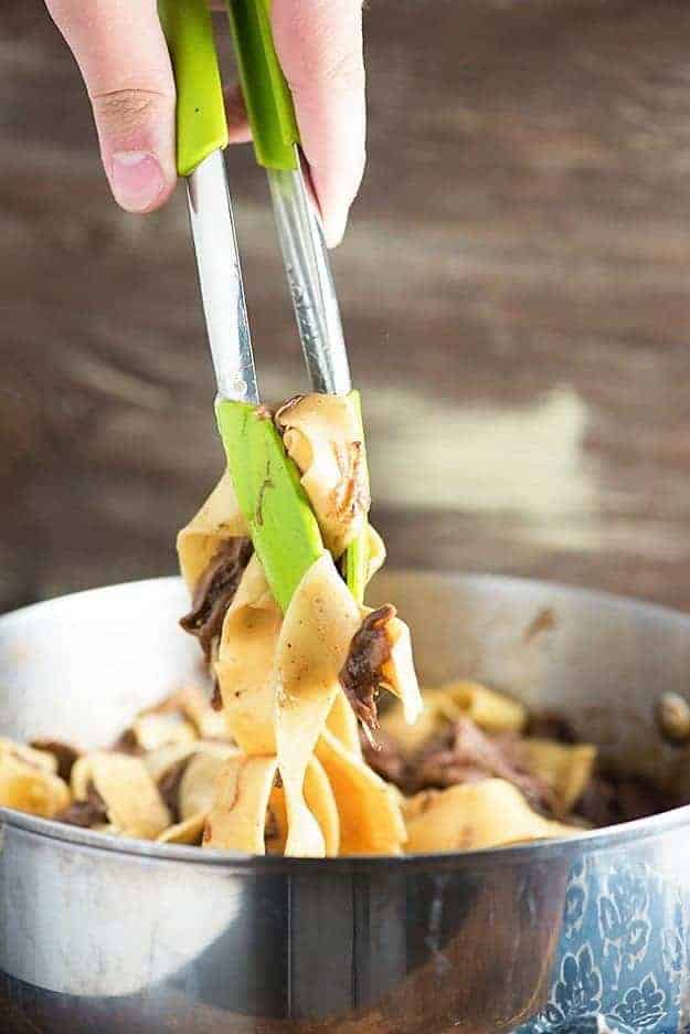 A set of tongs removing slow cooked beef and noodles from a pot.