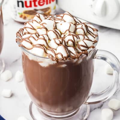 Mug full of hot chocolate in front of Nutella jar and crockpot.