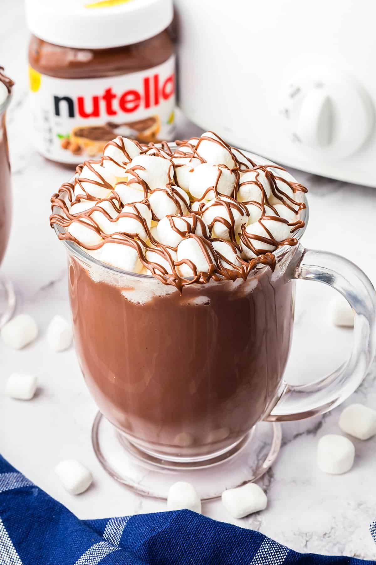 Mug full of hot chocolate in front of Nutella jar and crockpot.