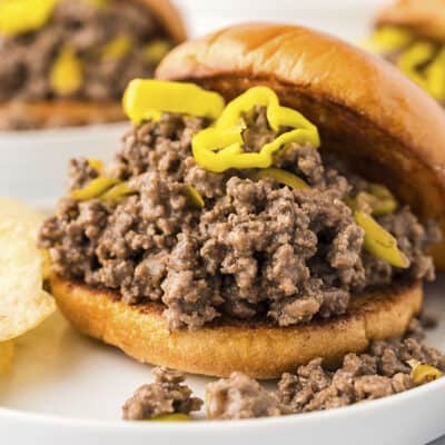 Sloppy joe sandwich topped with pepperoncini peppers on white plate with chips.