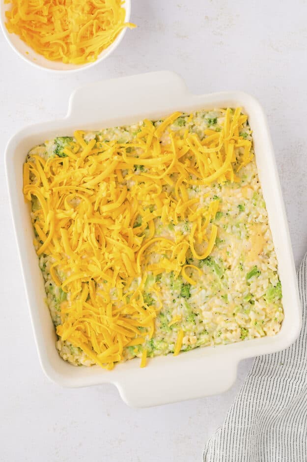 Broccoli rice casserole topped with shredded cheddar in white baking dish.