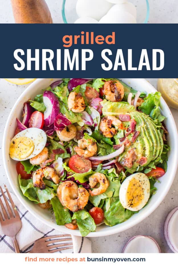 White bowl full of salad topped with grilled shrimp, tomatoes, and eggs.