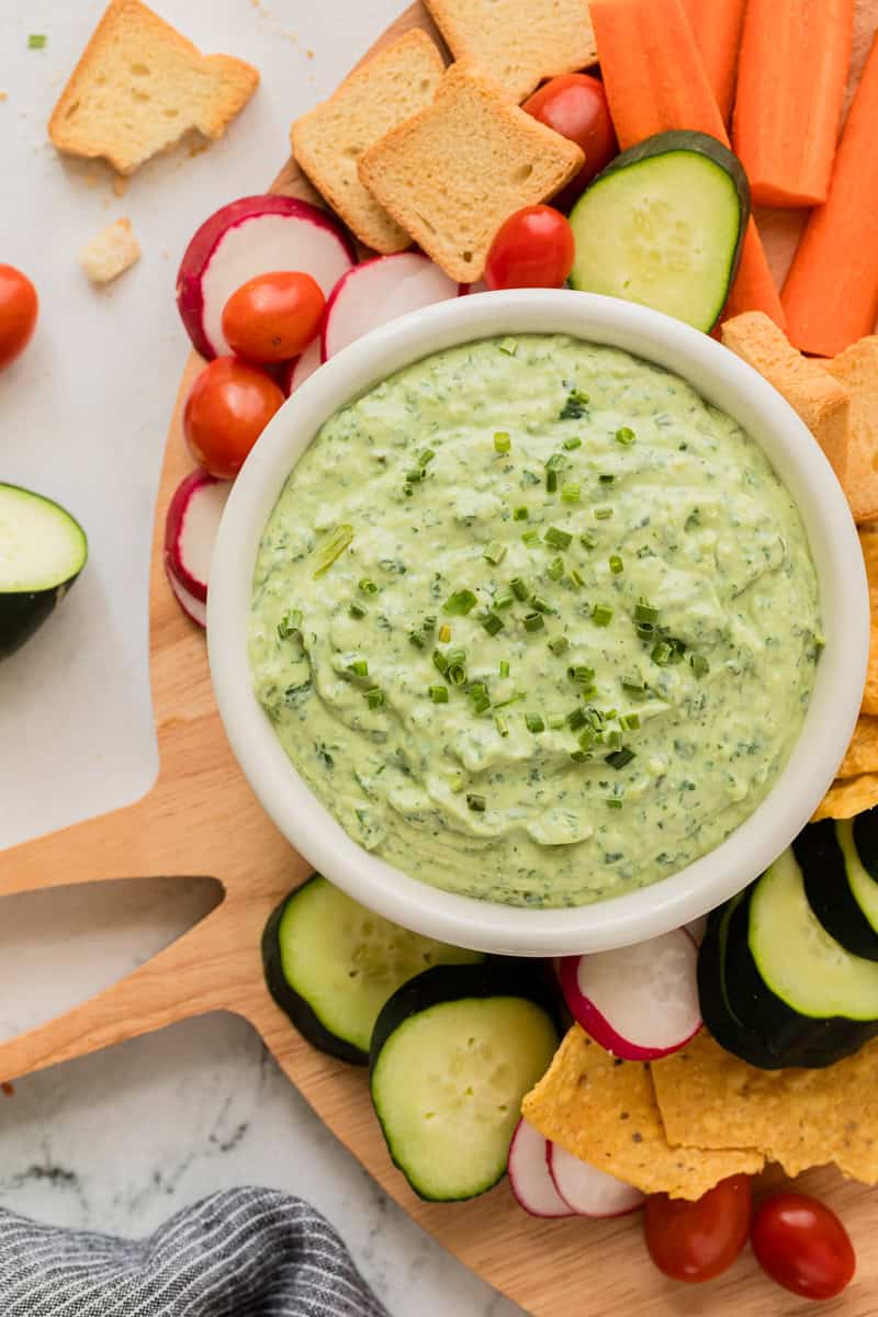 Green goddess dip in white bowl surrounded by fresh vegetables and crackers.