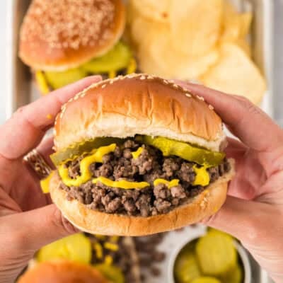 Hands holding a loose meat sandwich topped with pickles and mustard.