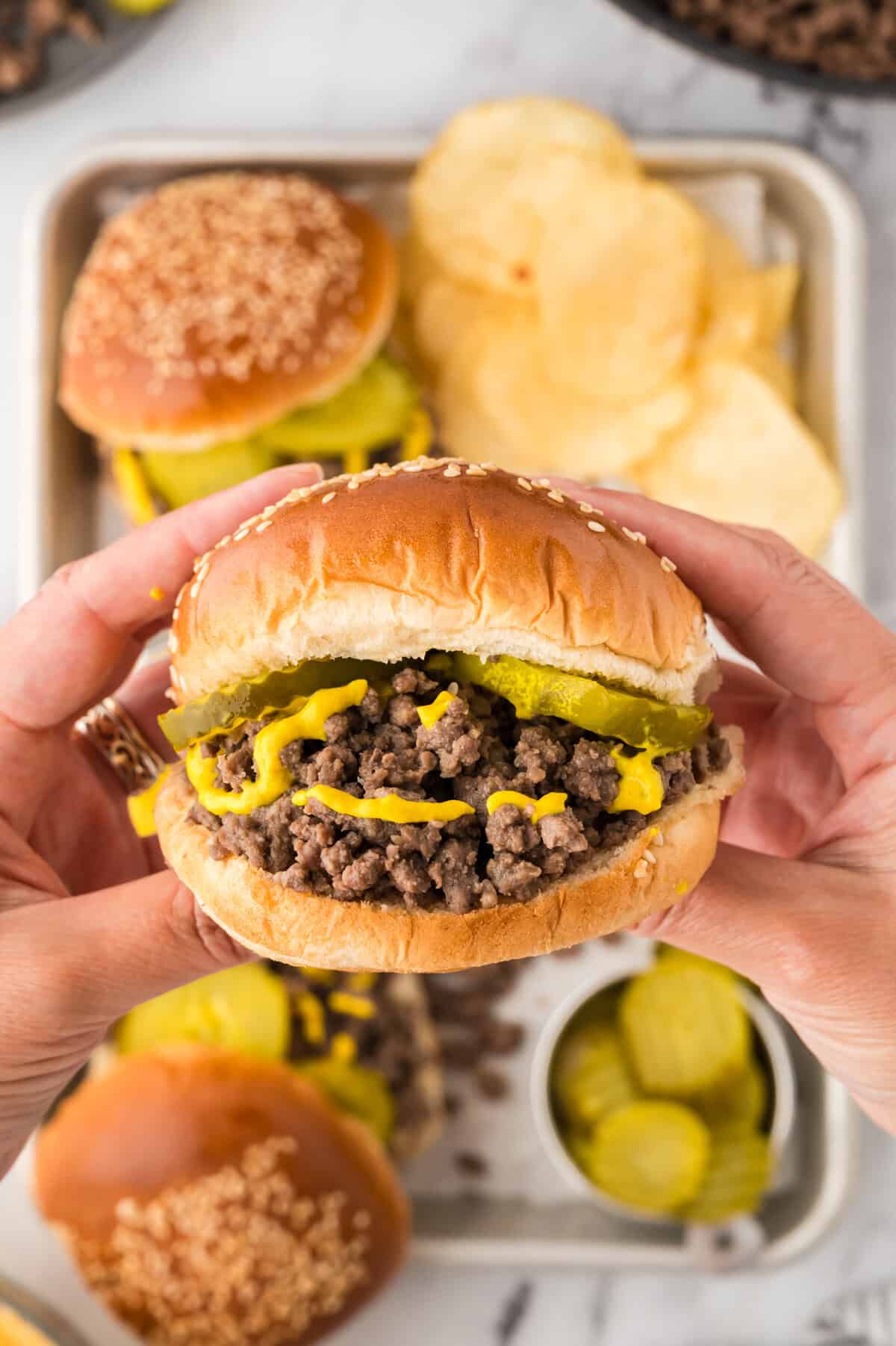 Hands holding a loose meat sandwich topped with pickles and mustard.