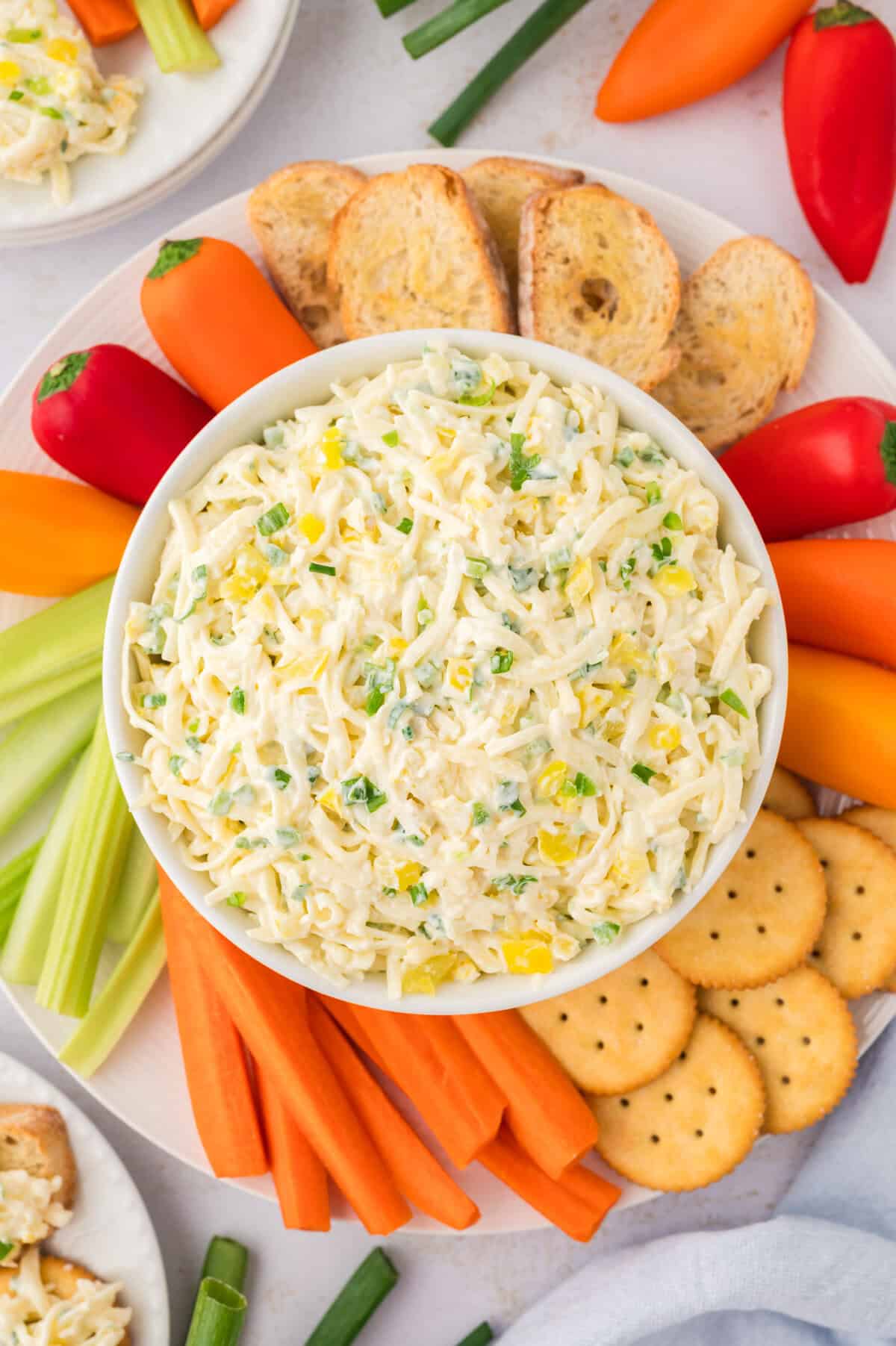 Bowl full of cheese slaw surrounded by crackers and vegetables.