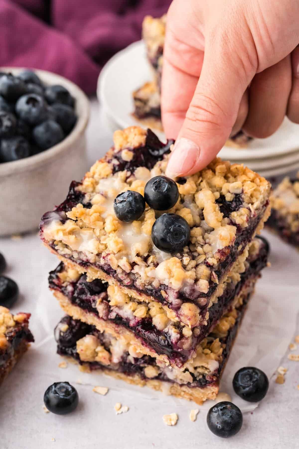 Hand grabbing a blueberry crumble bar off a stack.