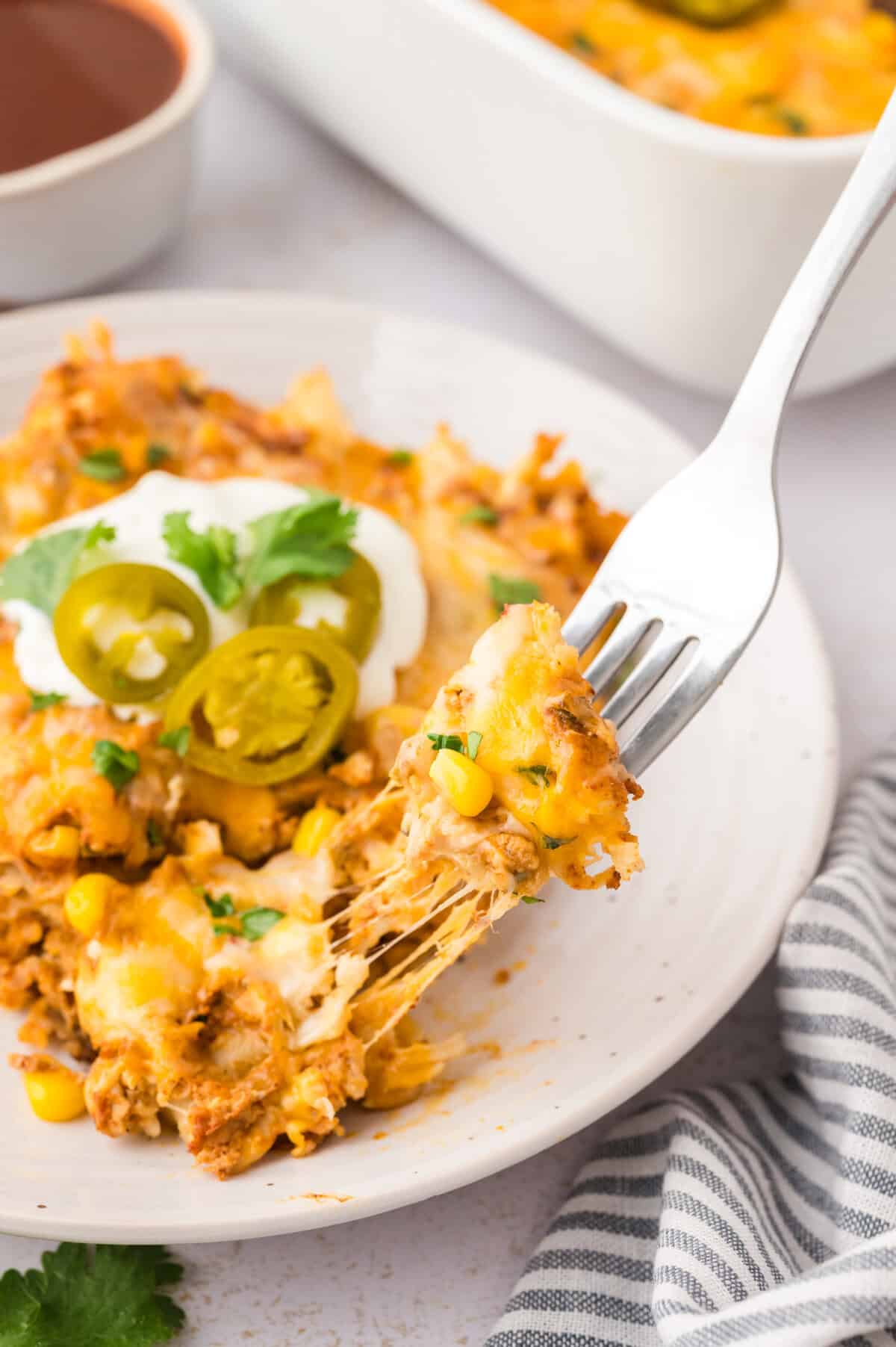 Fork taking a piece of sour cream chicken enchilada casserole off a white plate with cheese pull.