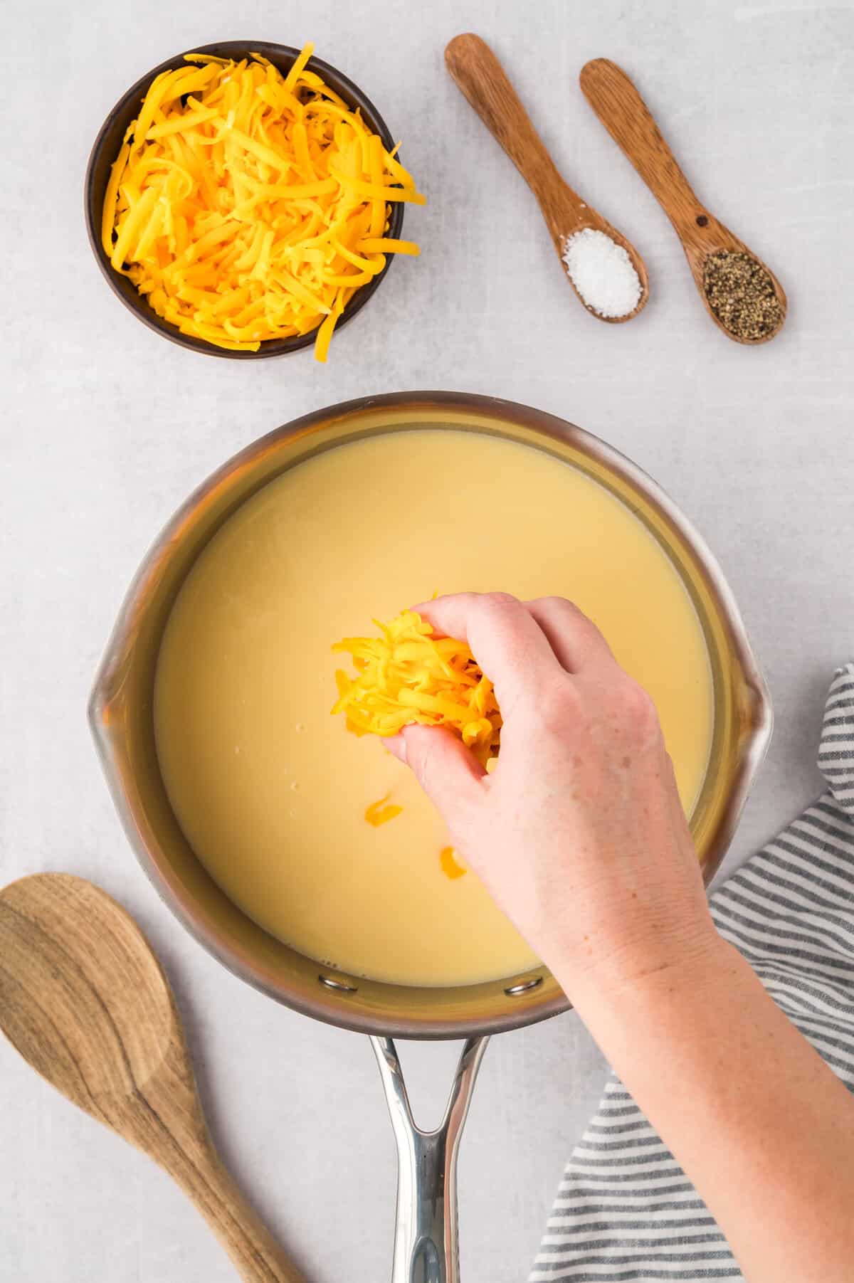 Hand dropping cheddar into cheese sauce recipe.